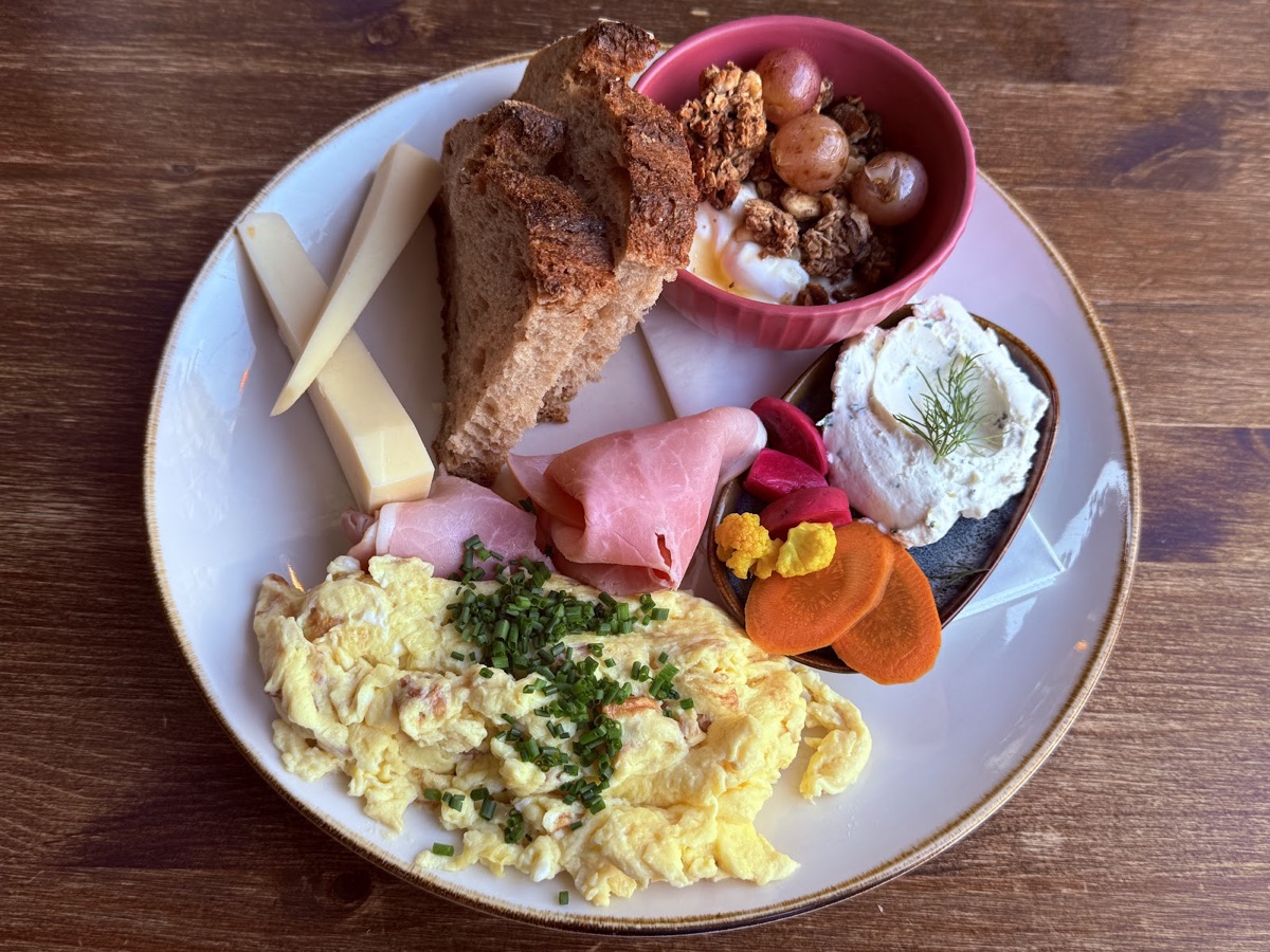 Frühstücksteller mit Eierspeis, Schinken, Käse, Brot, einer Schale Granola und Frischkäse