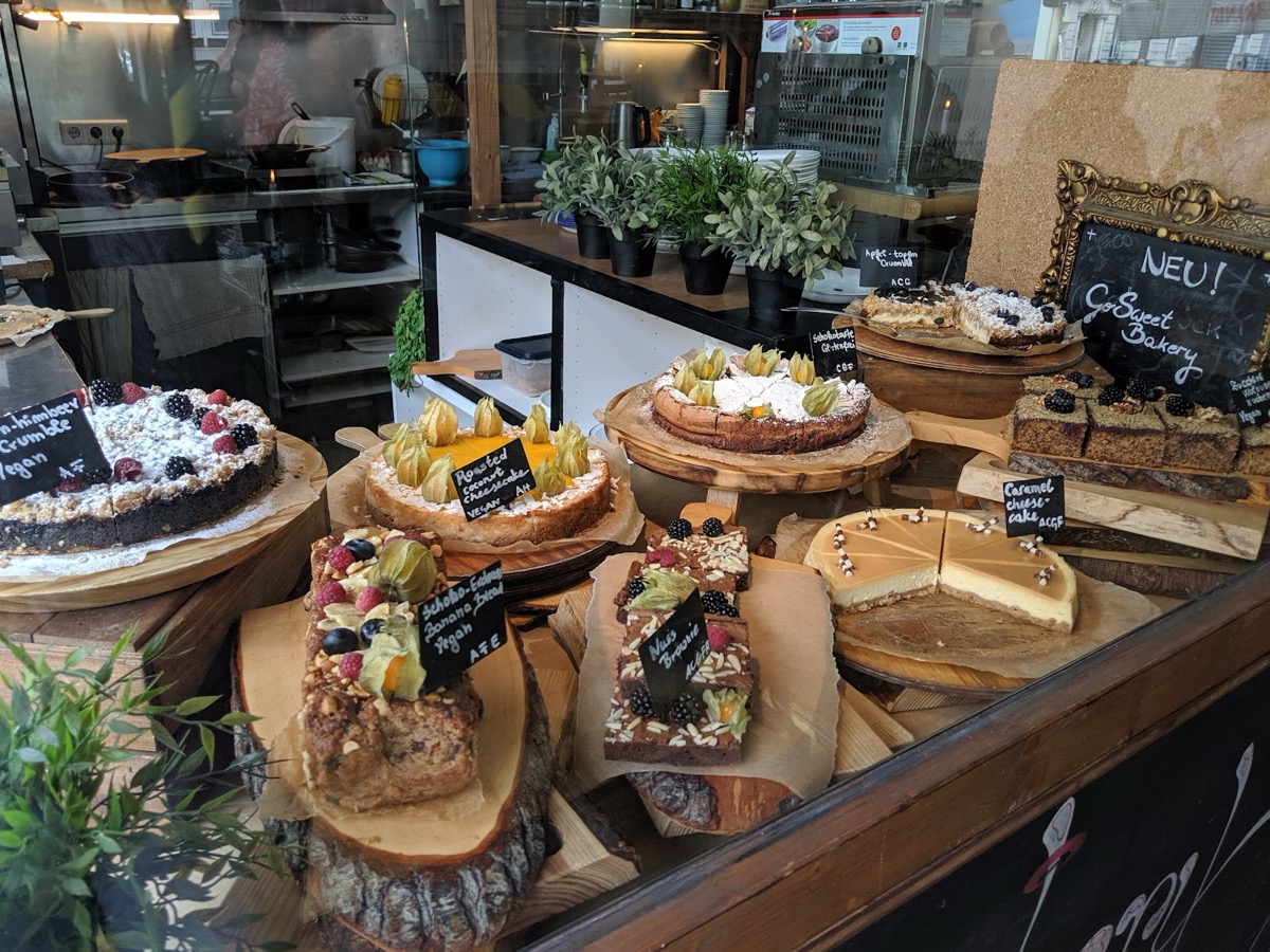 Kuchen auf Holzbrettern in der Auslage: Roasted Coconut Cheesecake (vegan), Schoko-Erdnuss-Banana-Bread, Nuss-Brownie und Karamell-Cheesecake